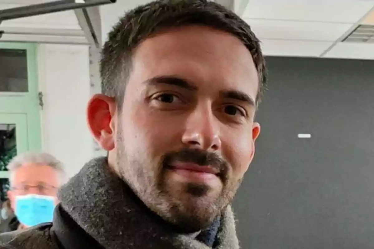 Hombre joven con barba y bufanda gris en un interior, al fondo una persona con mascarilla azul. Hombre joven con barba y bufanda gris en un interior, al fondo una persona con mascarilla azul.