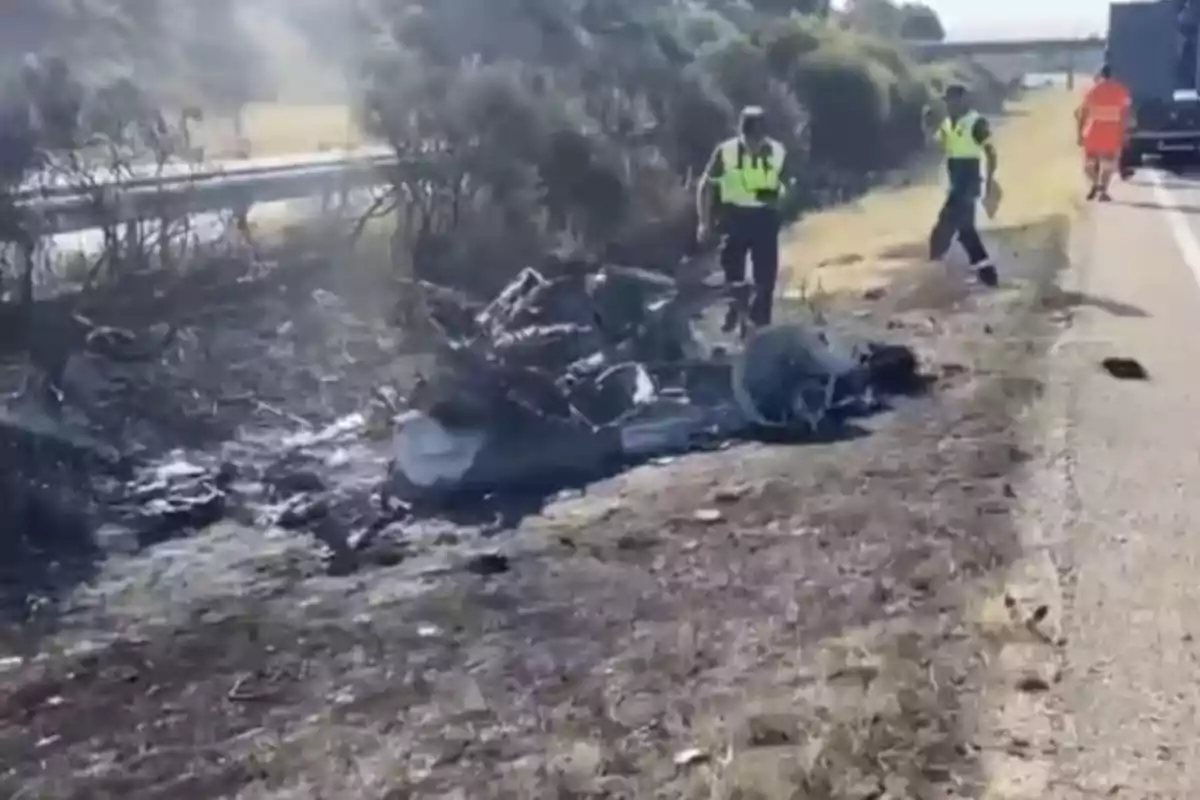 Charred remains of a vehicle on the side of a road with police and emergency personnel inspecting the scene. Charred remains of a vehicle on the side of a road with police and emergency personnel inspecting the scene.