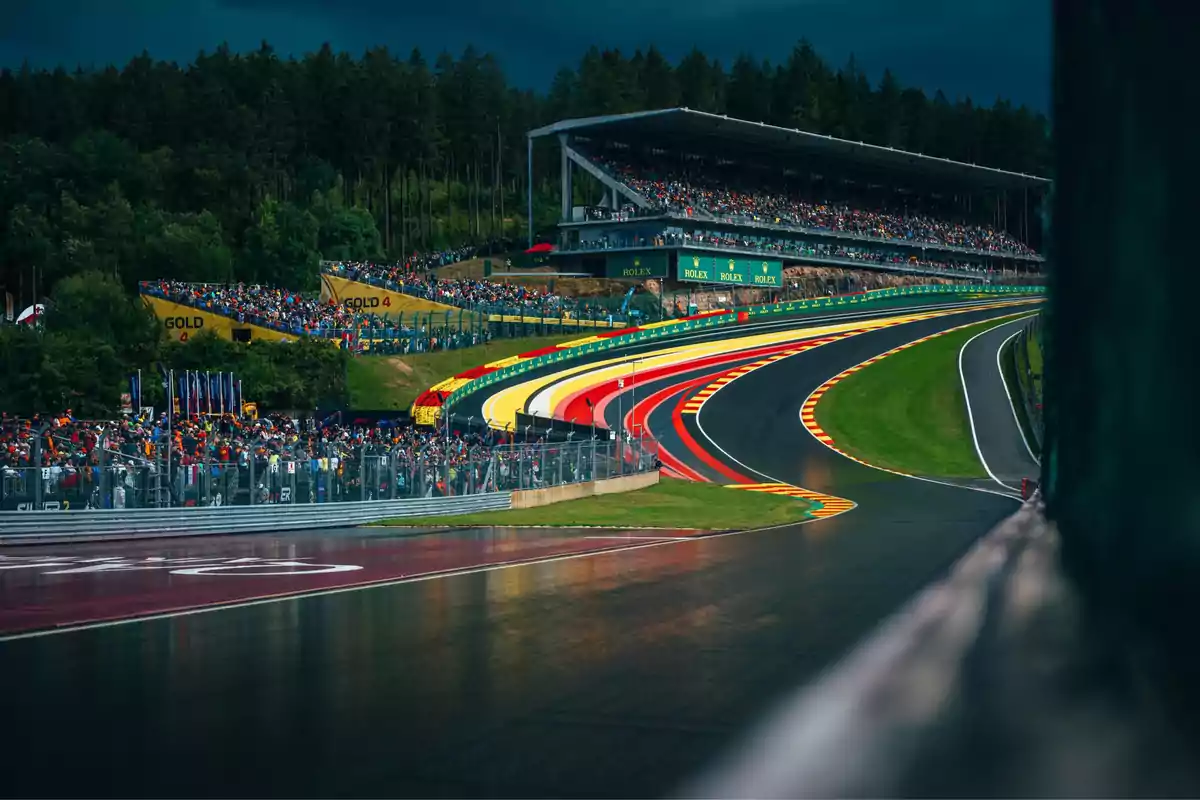 View of a racetrack with grandstands full of fans and a curve decorated with bright colors, surrounded by trees and a cloudy sky. View of a racetrack with grandstands full of fans and a curve decorated with bright colors, surrounded by trees and a cloudy sky.