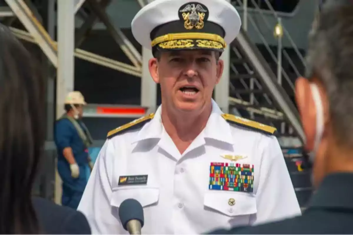 Un oficial naval con uniforme blanco y gorra está hablando frente a micrófonos mientras otras personas lo escuchan en un entorno industrial.