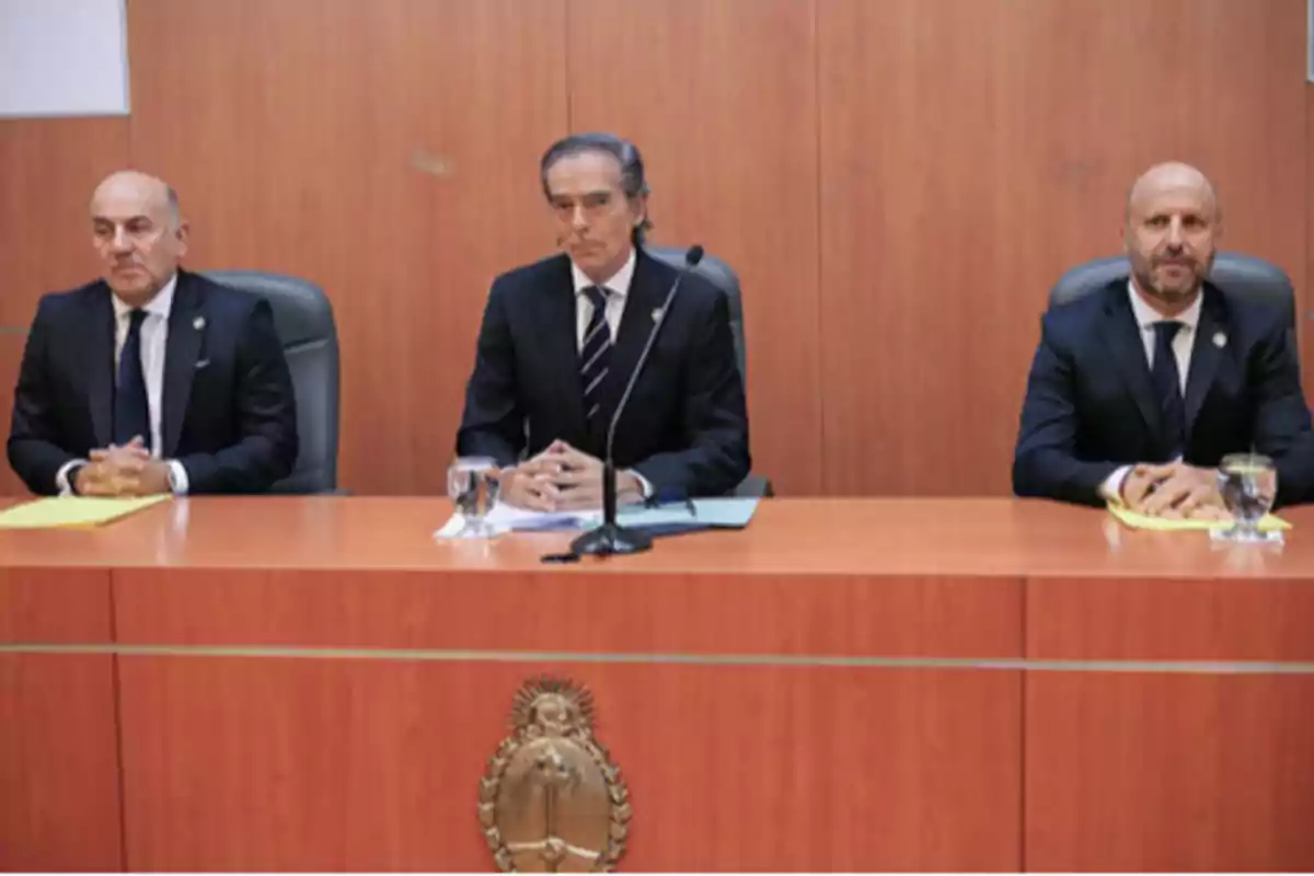 Tres hombres con traje oscuro sentados detrás de un estrado de madera en una sala formal con el escudo nacional de Argentina al frente Tres hombres con traje oscuro sentados detrás de un estrado de madera en una sala formal con el escudo nacional de Argentina al frente