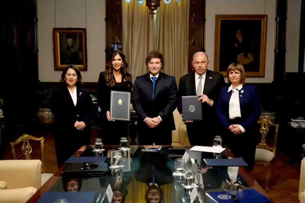 Cinco personas vestidas formalmente posan en una sala elegante con una mesa de reuniones al frente y cuadros en la pared de fondo