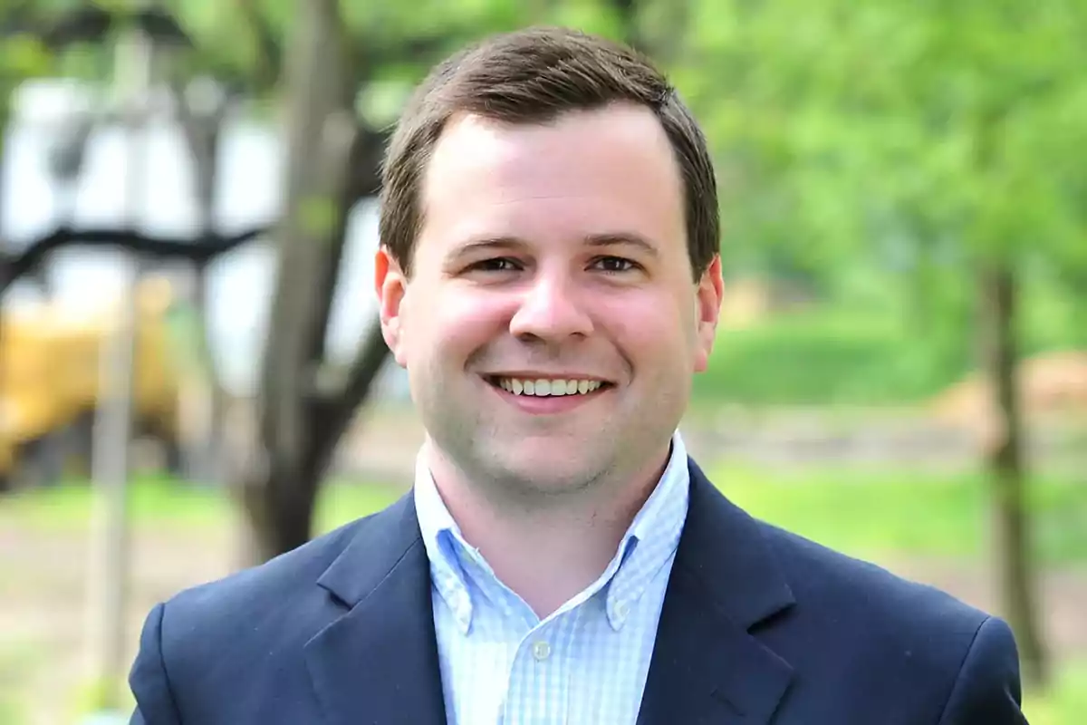 Young man with short dark hair smiling, wearing a blue jacket and light-colored jersey in an outdoor setting with green trees in the background Young man with short dark hair smiling, wearing a blue jacket and light-colored jersey in an outdoor setting with green trees in the background