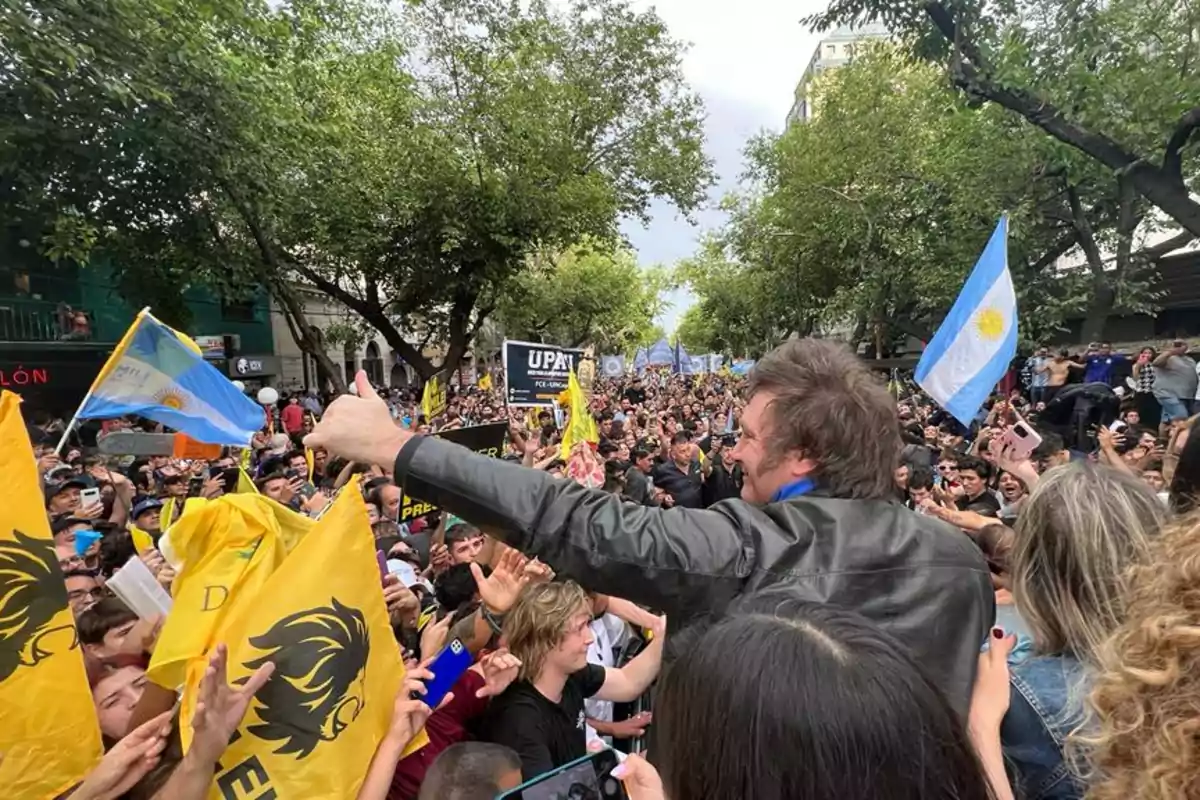 A man in a leather jacket greets a crowd on the street while several people hold Argentine flags and yellow flags with a black lion. A man in a leather jacket greets a crowd on the street while several people hold Argentine flags and yellow flags with a black lion.