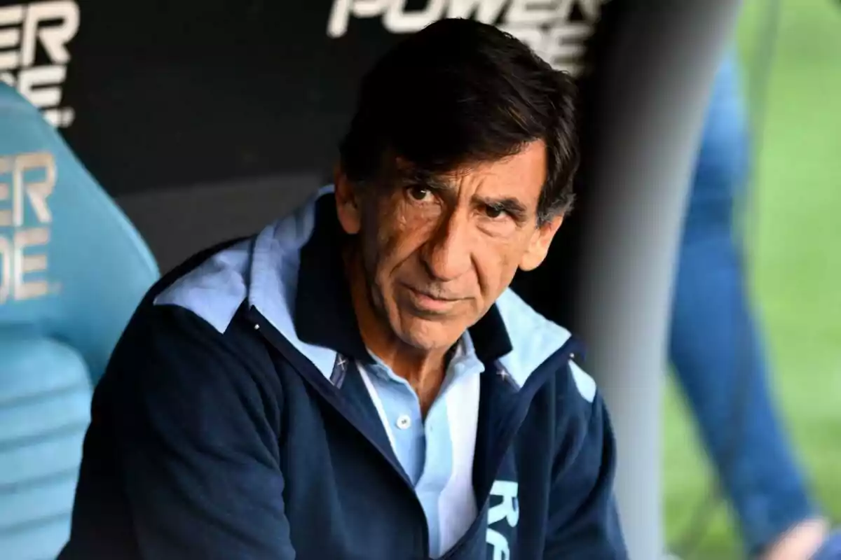 Dark-haired man with a serious expression sitting on a sports bench wearing a blue jacket
