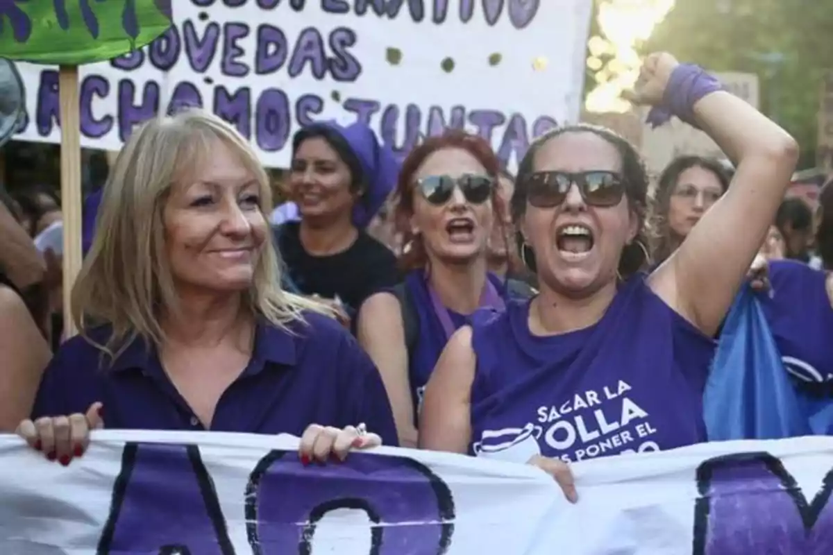 Women participating in a feminist demonstration wearing purple jerseys and holding purple signs while showing expressions of joy and determination