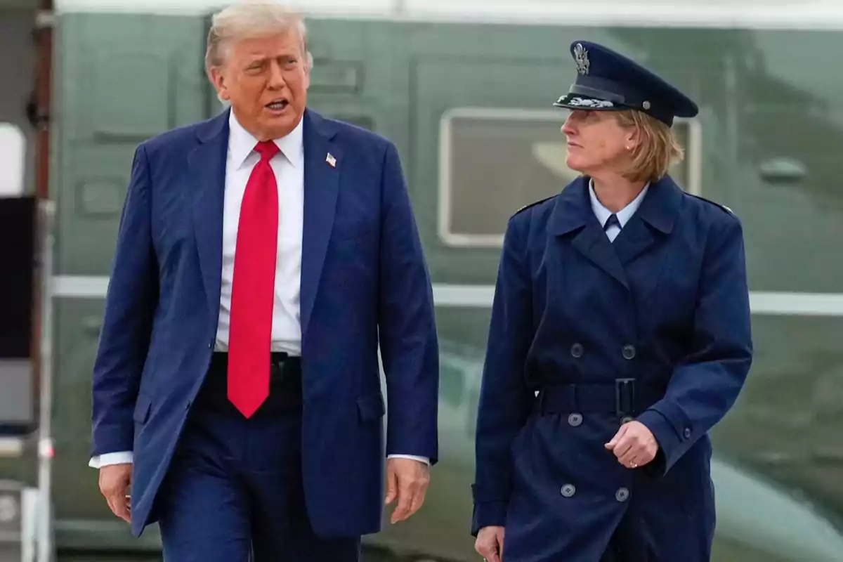 A man in a blue suit and red tie walks next to a woman in a blue military uniform in front of an official vehicle. A man in a blue suit and red tie walks next to a woman in a blue military uniform in front of an official vehicle.
