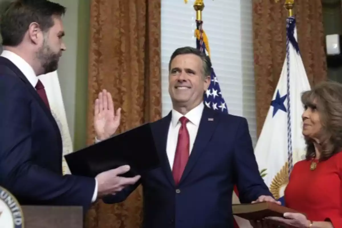 A man in a blue suit and red tie raises his right hand while another person holds a book and a woman in a red dress watches him, in a formal setting with flags in the background. A man in a blue suit and red tie raises his right hand while another person holds a book and a woman in a red dress watches him, in a formal setting with flags in the background.