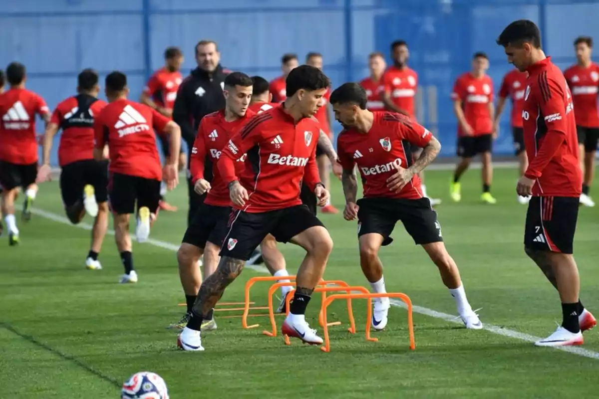 Jugadores de fútbol entrenando en una cancha de césped con vallas naranjas y usando camisetas rojas