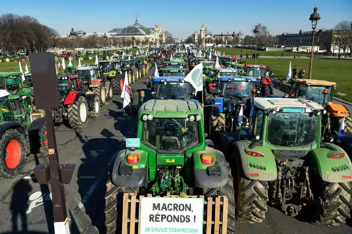 Fila de tractores ocupando una avenida en una manifestación agrícola en París con un cartel en el tractor del frente que pide respuesta al presidente Macron Fila de tractores ocupando una avenida en una manifestación agrícola en París con un cartel en el tractor del frente que pide respuesta al presidente Macron