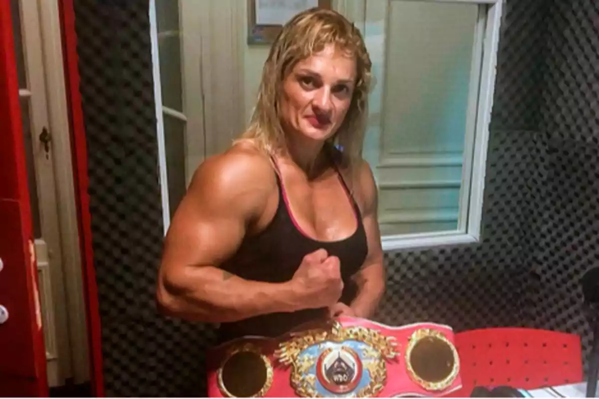 Mujer musculosa con camiseta deportiva negra posando con un cinturón de campeonato de boxeo en una sala con paredes acolchadas.