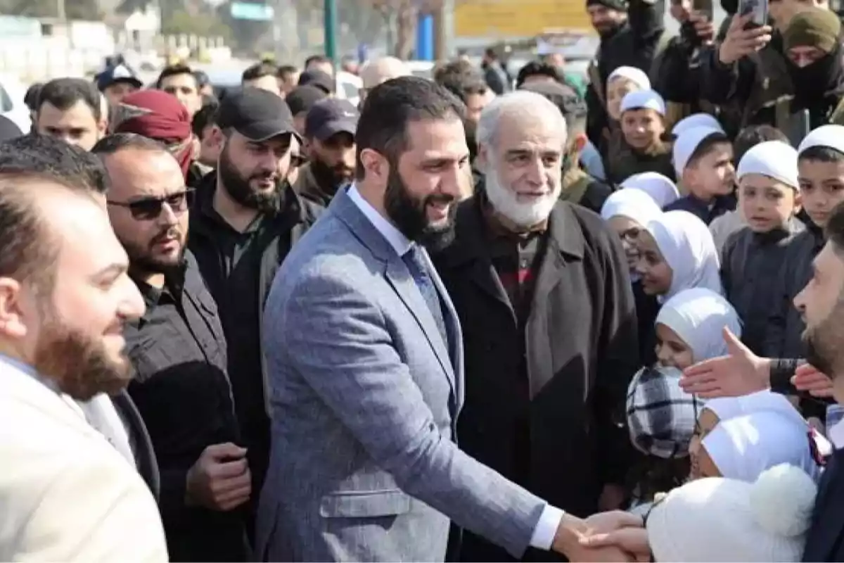 Un hombre con traje saluda a un grupo de personas en un evento al aire libre rodeado de niños y adultos Un hombre con traje saluda a un grupo de personas en un evento al aire libre rodeado de niños y adultos