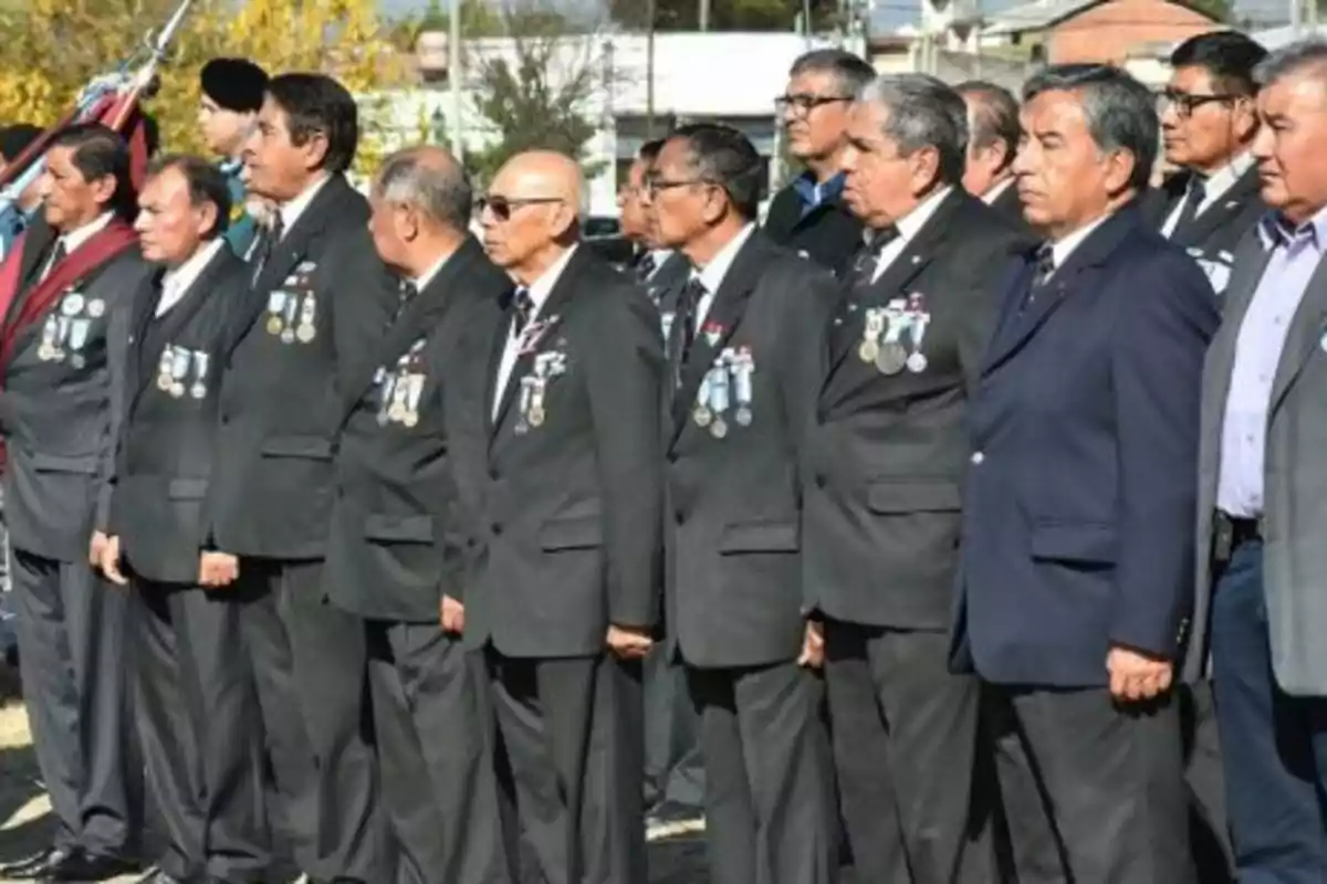 A group of elderly men dressed in dark suits with medals on their chests are lined up during an outdoor ceremony. A group of elderly men dressed in dark suits with medals on their chests are lined up during an outdoor ceremony.