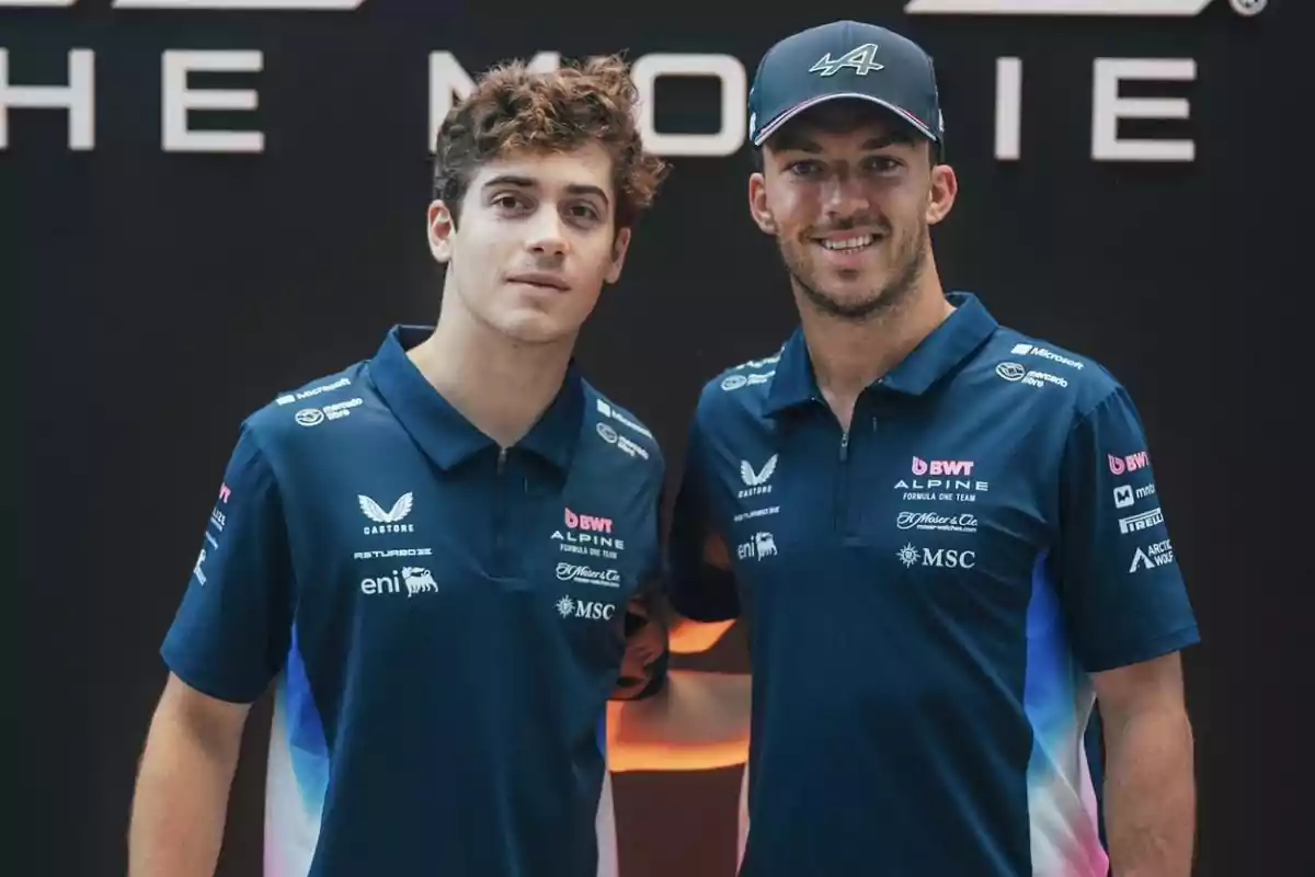 Dos hombres con uniforme azul del equipo Alpine de Fórmula 1 posan juntos y sonríen frente a un fondo oscuro Dos hombres con uniforme azul del equipo Alpine de Fórmula 1 posan juntos y sonríen frente a un fondo oscuro
