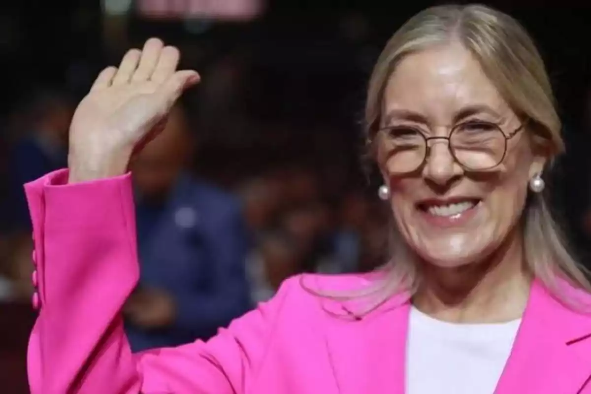 Mujer de cabello rubio y largo con gafas y saco rosa saludando y sonriendo