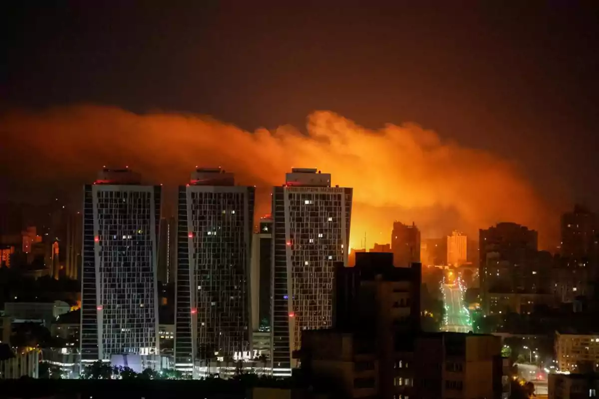 Vista nocturna de una ciudad con varios edificios altos en primer plano y una gran nube naranja iluminada en el cielo al fondo, sugiriendo un incendio o explosión