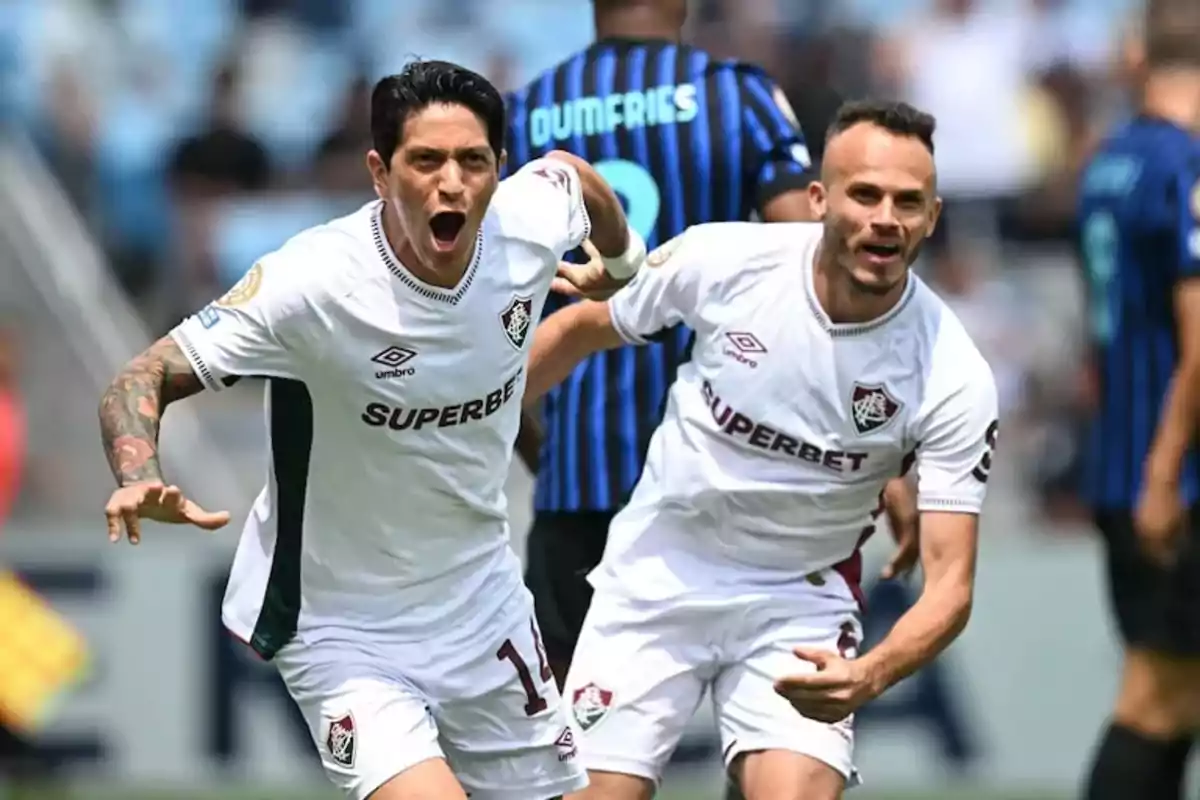 Dos futbolistas del Fluminense celebran un gol durante un partido, ambos con uniforme blanco y expresión de alegría, mientras jugadores rivales aparecen al fondo. Dos futbolistas del Fluminense celebran un gol durante un partido, ambos con uniforme blanco y expresión de alegría, mientras jugadores rivales aparecen al fondo.