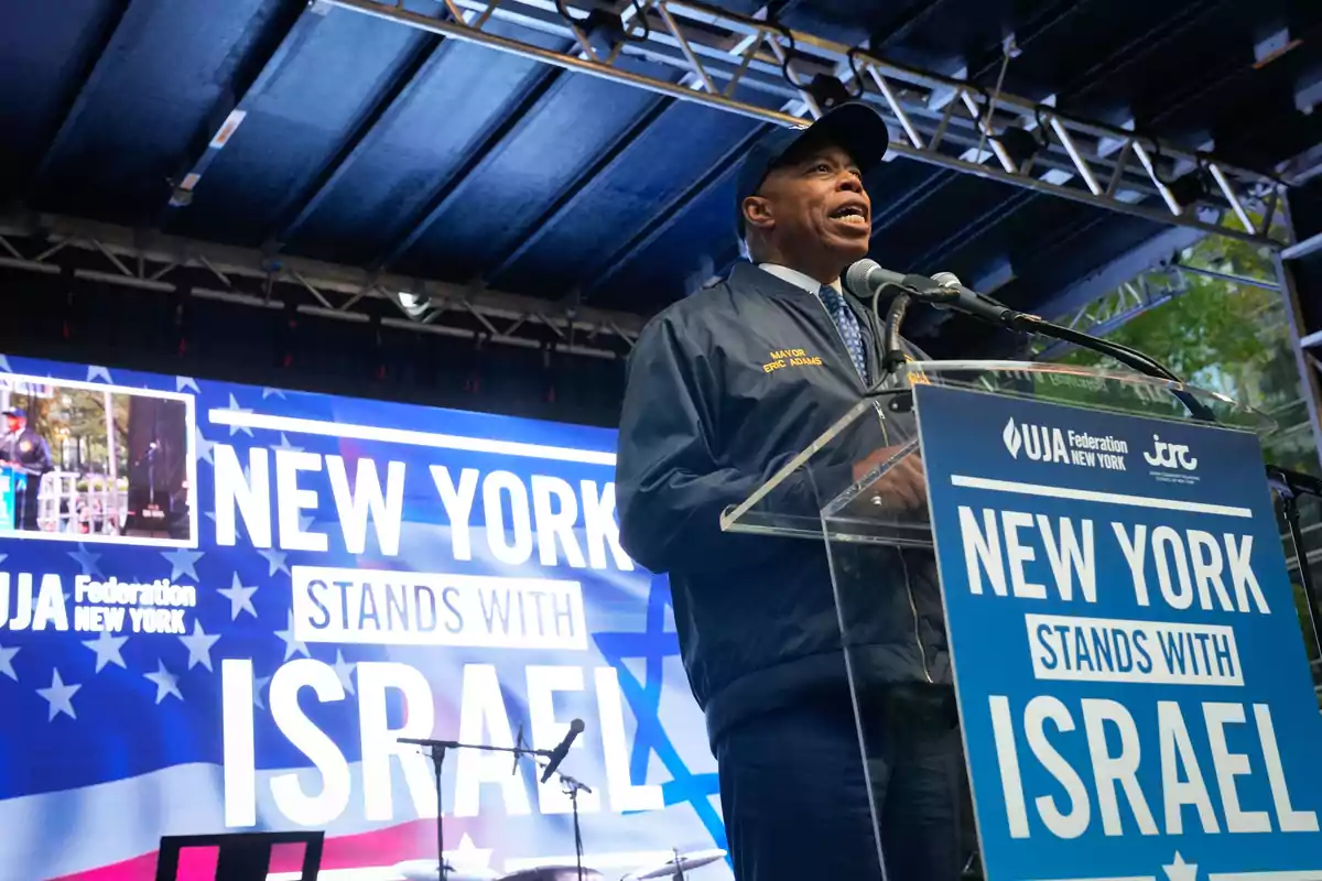 A man wearing a jacket and a cap speaks on a stage in front of a microphone, with a sign and a screen in the background that say A man wearing a jacket and a cap speaks on a stage in front of a microphone, with a sign and a screen in the background that say
