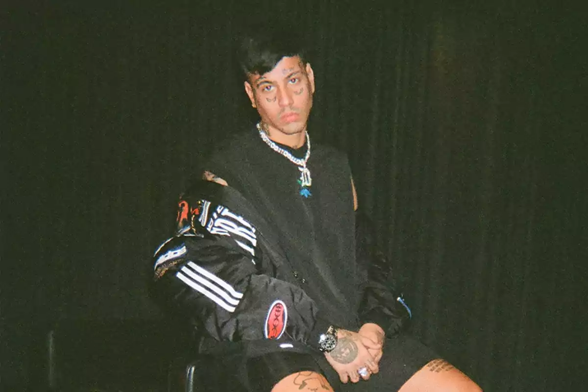 Persona joven con tatuajes en la cara y las manos, vistiendo ropa oscura y accesorios llamativos, sentada frente a un fondo negro