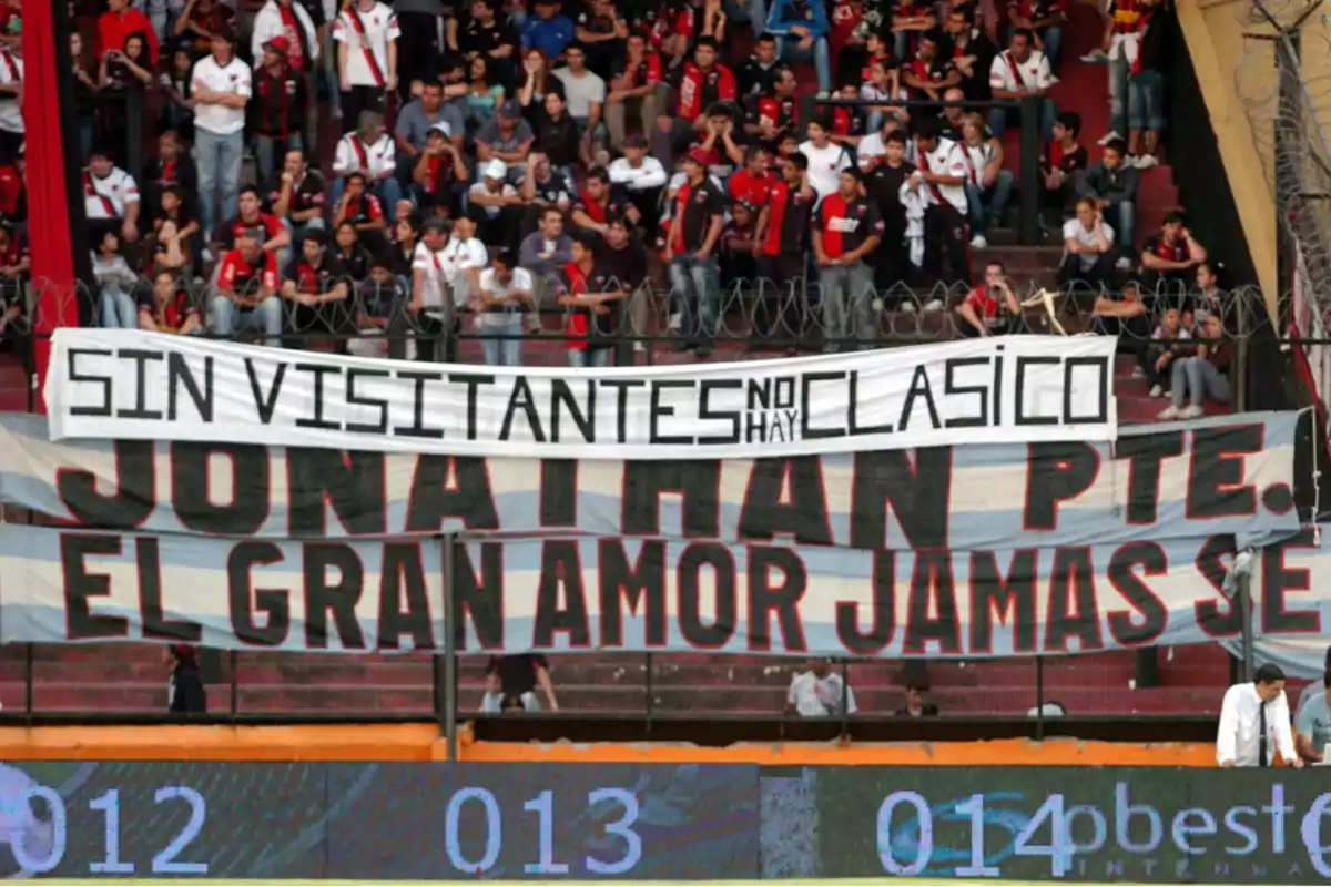 Fans in a stadium stand with flags displaying messages supporting their team and protesting the absence of visiting supporters.