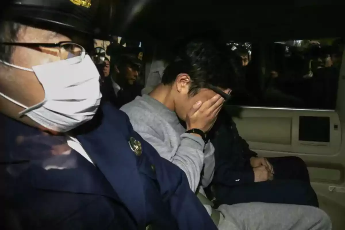 Un hombre cubre su rostro con la mano mientras es escoltado por un oficial de policía dentro de un vehículo Un hombre cubre su rostro con la mano mientras es escoltado por un oficial de policía dentro de un vehículo
