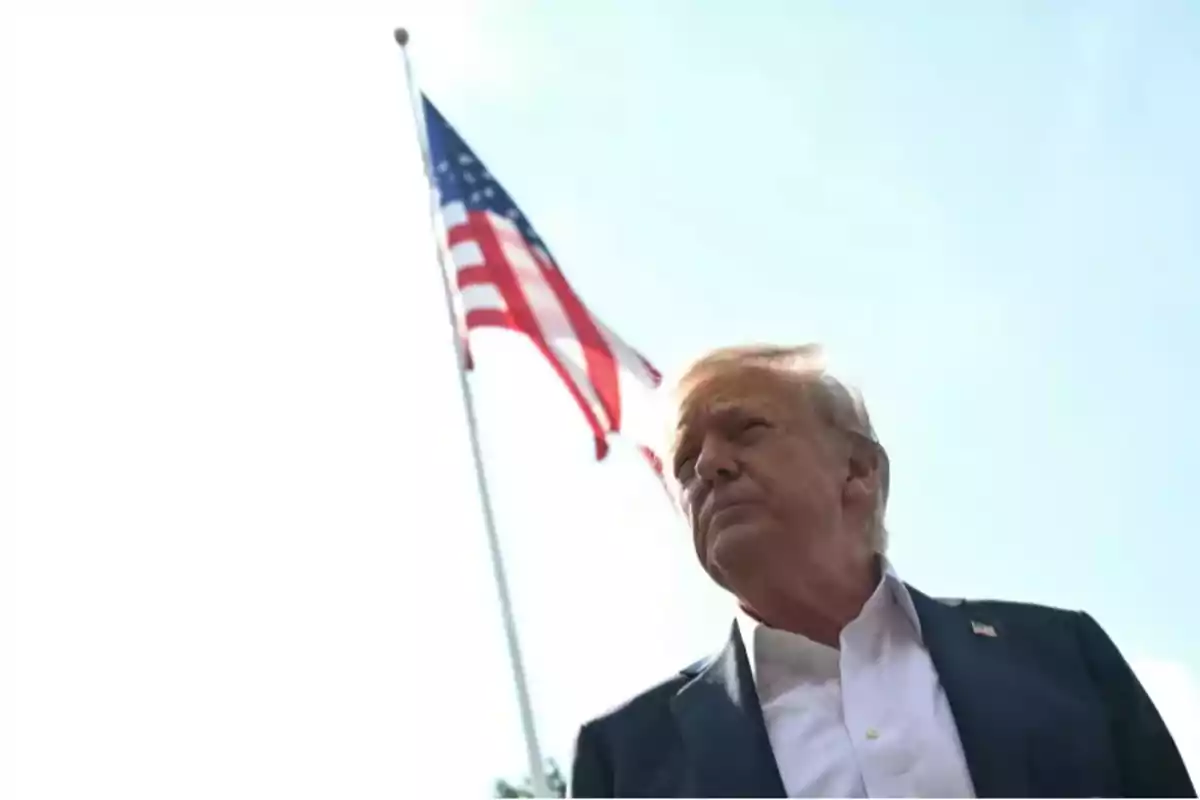 Hombre de cabello rubio con traje oscuro y camisa blanca de pie frente a una bandera de Estados Unidos ondeando en el fondo