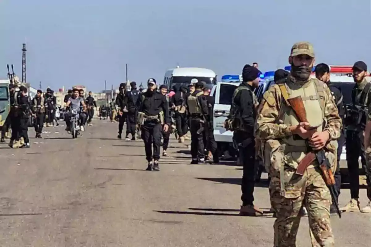 Grupo de personas armadas con uniformes militares y vehículos estacionados en una carretera bajo un cielo despejado Grupo de personas armadas con uniformes militares y vehículos estacionados en una carretera bajo un cielo despejado