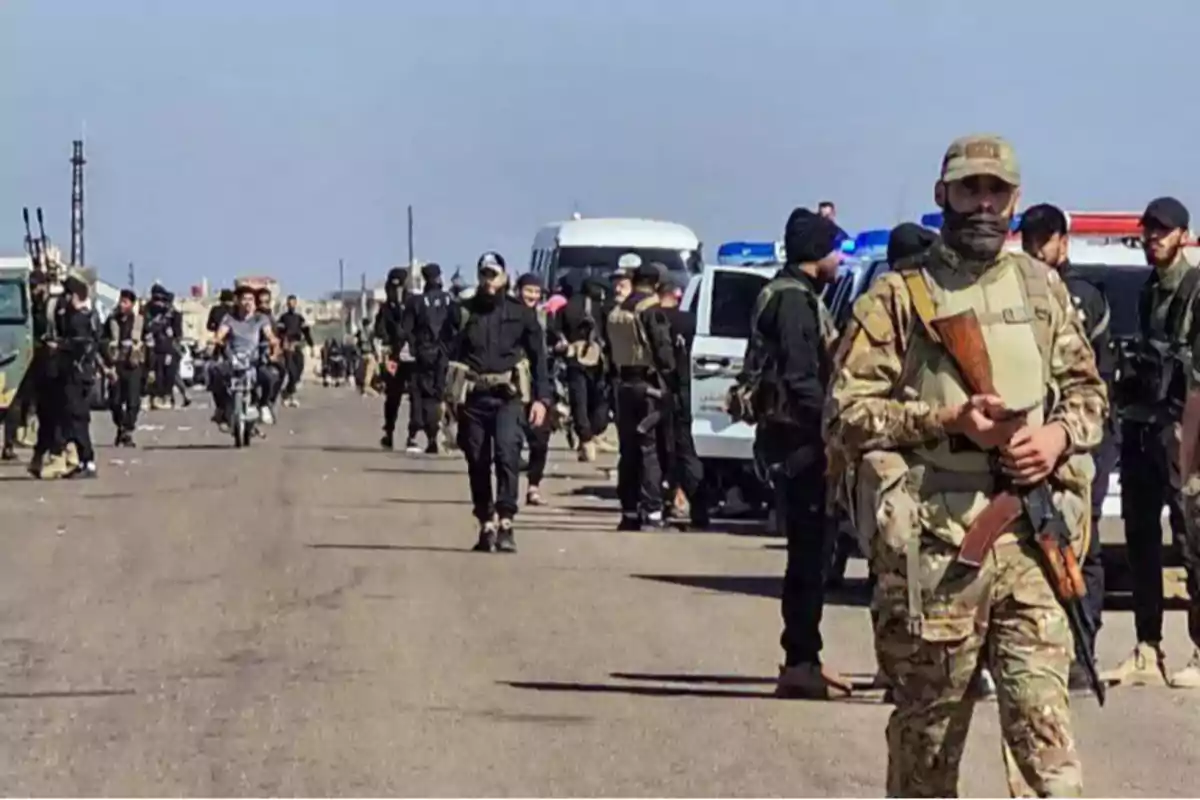 Grupo de personas armadas con uniformes militares y vehículos estacionados en una carretera bajo un cielo despejado