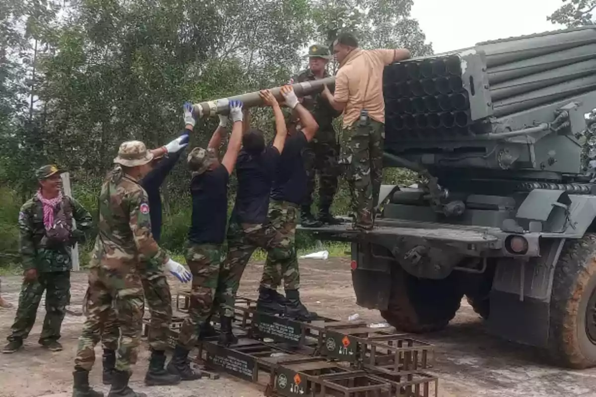 Soldiers in camouflage uniforms load a large shell into a multiple rocket launcher mounted on a truck in an outdoor setting surrounded by trees.