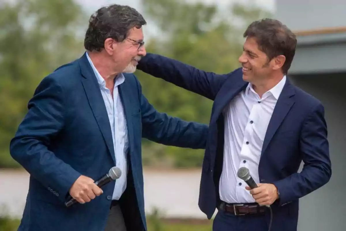 Dos hombres con traje sonríen y se saludan mientras sostienen micrófonos al aire libre Dos hombres con traje sonríen y se saludan mientras sostienen micrófonos al aire libre