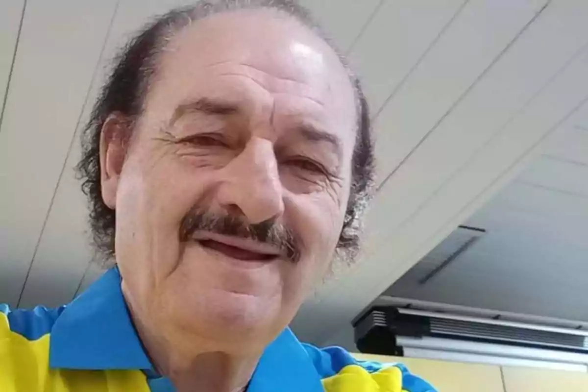 Hombre de mediana edad con bigote y cabello rizado usando una camisa azul y amarilla sonríe en un ambiente interior iluminado Hombre de mediana edad con bigote y cabello rizado usando una camisa azul y amarilla sonríe en un ambiente interior iluminado
