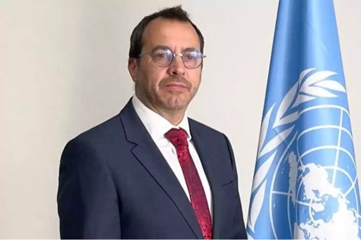 Hombre con gafas y traje oscuro posando junto a una bandera azul de la ONU