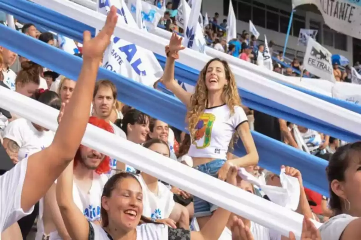 Personas celebrando en una tribuna con banderas y tiras de tela blancas y celestes, mostrando alegría y entusiasmo. Personas celebrando en una tribuna con banderas y tiras de tela blancas y celestes, mostrando alegría y entusiasmo.