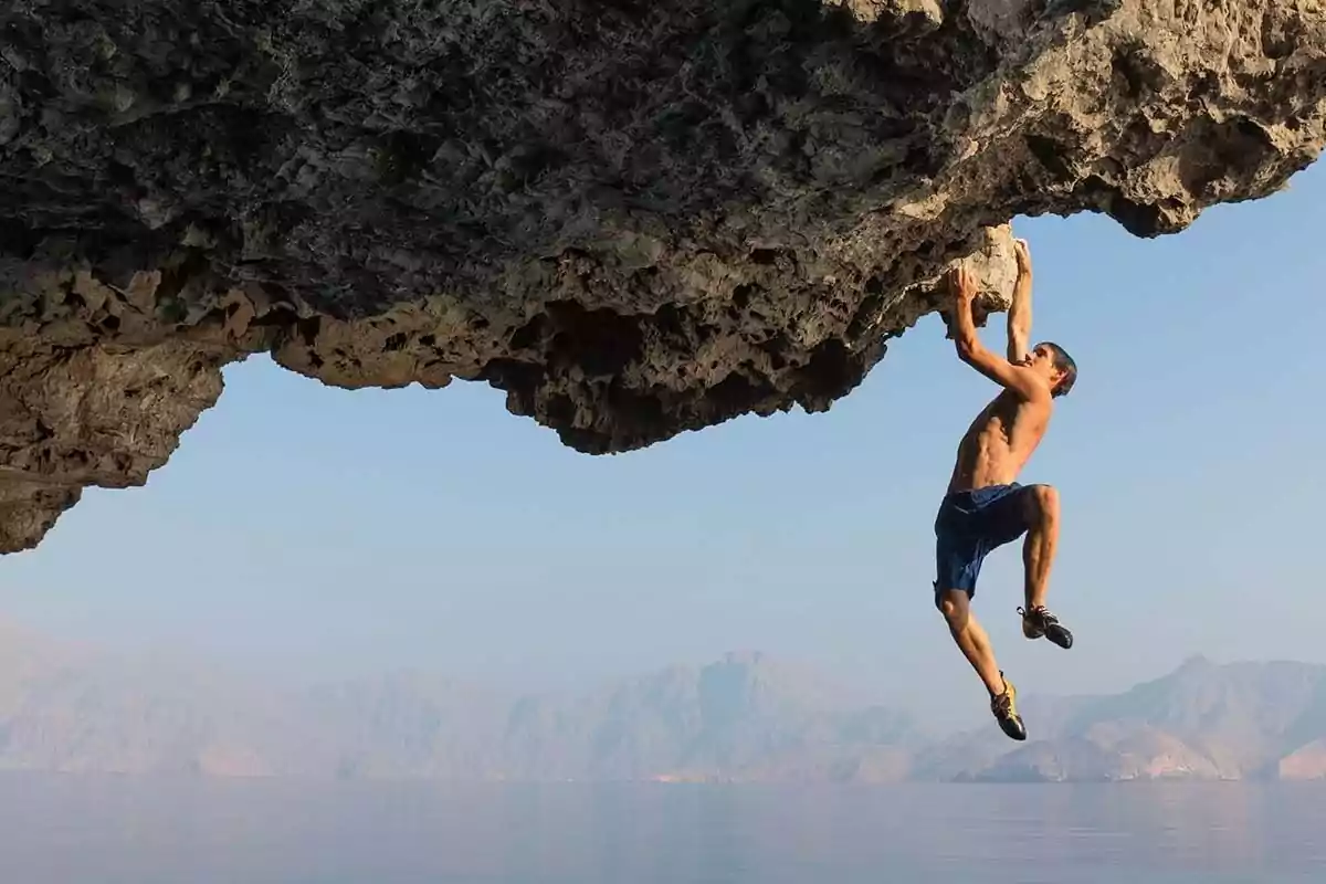 Hombre escalando una roca sobre el mar con montañas al fondo en un día despejado