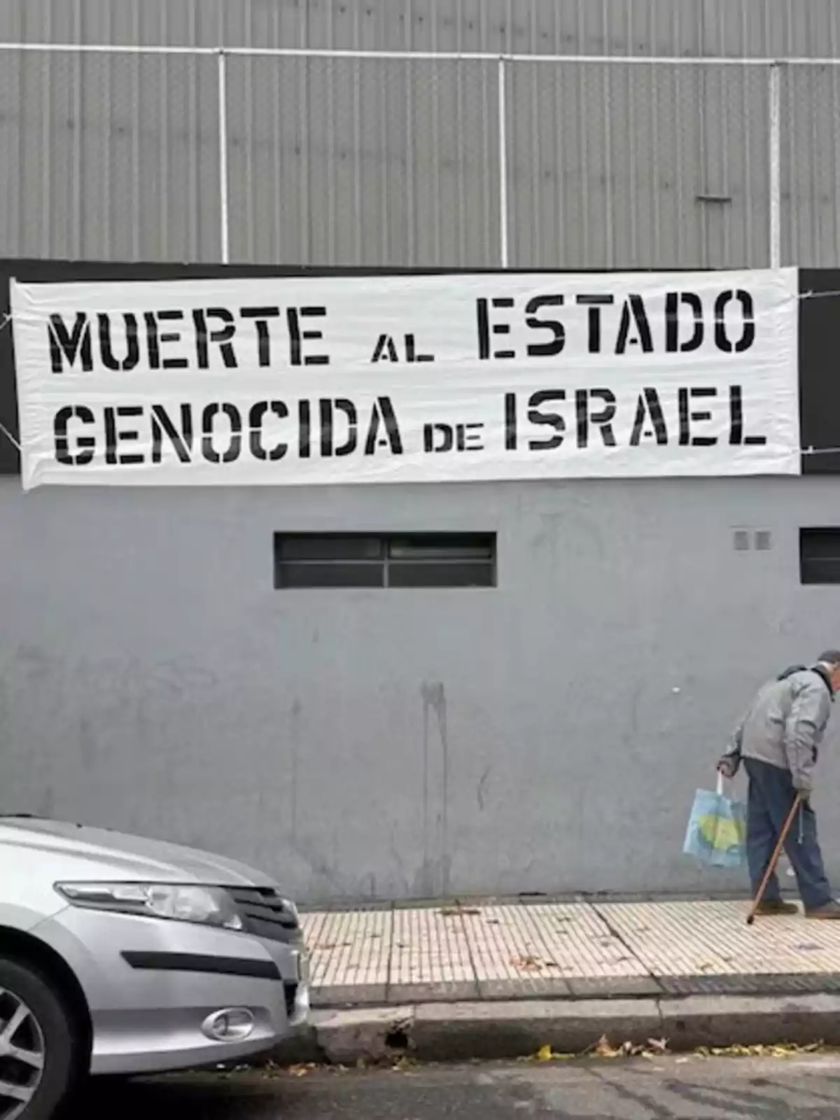 Banner with a strong political message hanging on a gray wall, an elderly man walks with a cane and carries a bag, while a silver car is parked on the street. Banner with a strong political message hanging on a gray wall, an elderly man walks with a cane and carries a bag, while a silver car is parked on the street.