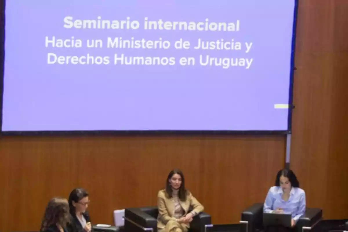 Four women seated in black armchairs participate in a panel in front of a screen displaying the title International Seminar: Towards a Ministry of Justice and Human Rights in Uruguay. Four women seated in black armchairs participate in a panel in front of a screen displaying the title International Seminar: Towards a Ministry of Justice and Human Rights in Uruguay.
