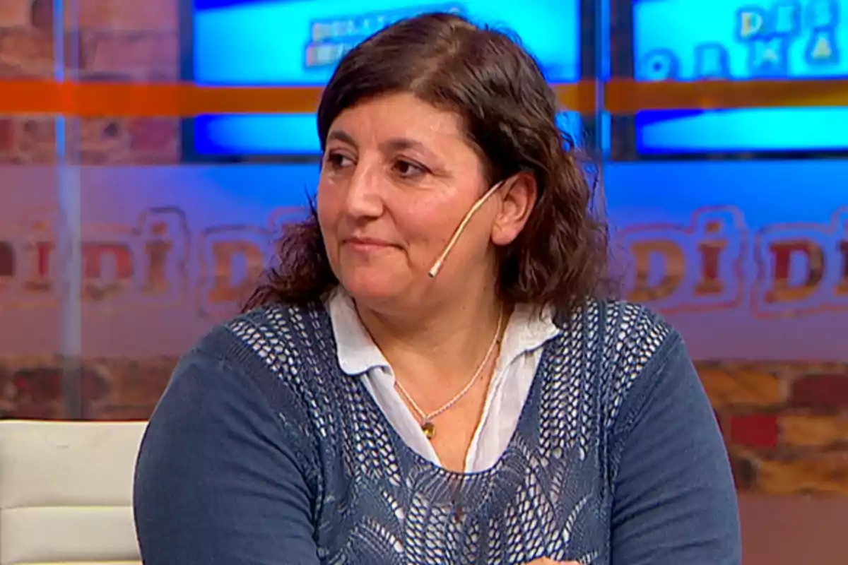 Mujer de cabello castaño y ondulado sentada en un estudio de televisión con fondo azul y letras desenfocadas, lleva un micrófono de diadema y viste un suéter azul sobre una camisa blanca Mujer de cabello castaño y ondulado sentada en un estudio de televisión con fondo azul y letras desenfocadas, lleva un micrófono de diadema y viste un suéter azul sobre una camisa blanca