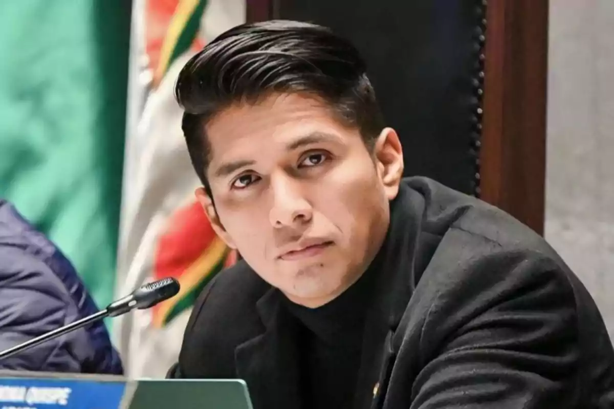 Young man with dark hair styled to the side is wearing a black jacket and is seated in front of a microphone with a flag in the background.