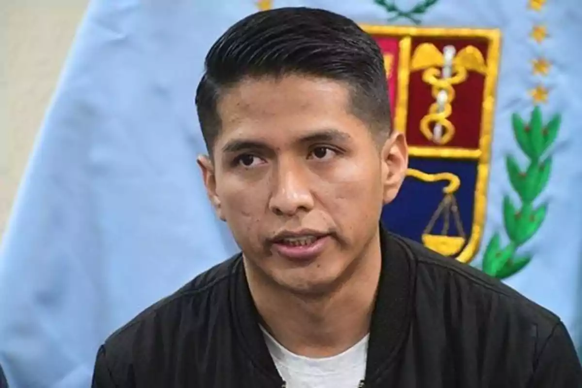 Un hombre joven de cabello corto y oscuro viste una chaqueta negra y está frente a un fondo azul claro con un escudo bordado Un hombre joven de cabello corto y oscuro viste una chaqueta negra y está frente a un fondo azul claro con un escudo bordado