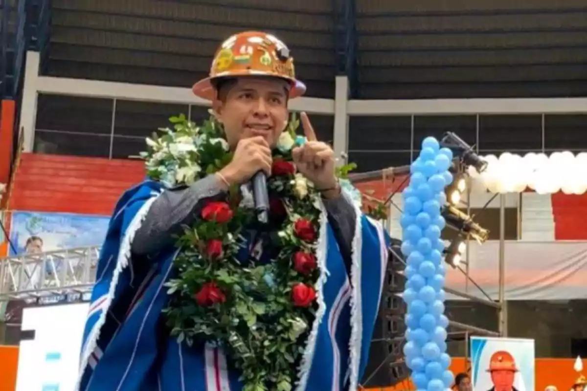 Hombre con casco de minero y poncho azul hablando en un micrófono, lleva una guirnalda de flores y está en un evento decorado con globos azules