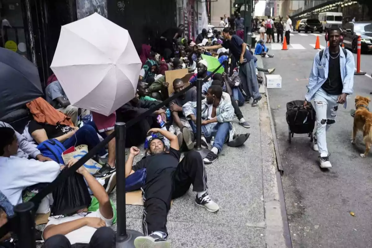 Varias personas descansan y esperan sentadas o acostadas en una acera junto a una calle concurrida, algunas con paraguas para protegerse del sol, mientras un hombre camina con una maleta y un perro. Varias personas descansan y esperan sentadas o acostadas en una acera junto a una calle concurrida, algunas con paraguas para protegerse del sol, mientras un hombre camina con una maleta y un perro.
