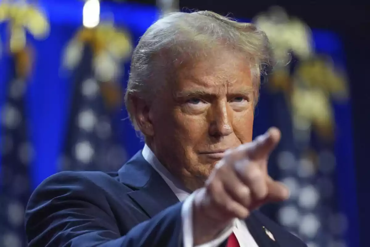 Hombre de cabello rubio con traje azul señalando hacia el frente con expresión seria y fondo de banderas desenfocadas Hombre de cabello rubio con traje azul señalando hacia el frente con expresión seria y fondo de banderas desenfocadas