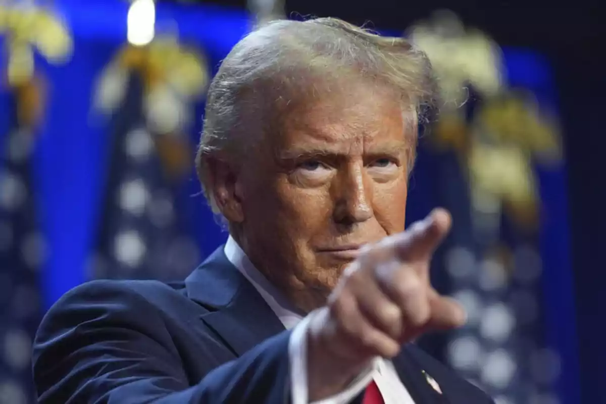 Blond-haired man in a blue suit pointing forward with a serious expression and a background of blurred flags Blond-haired man in a blue suit pointing forward with a serious expression and a background of blurred flags