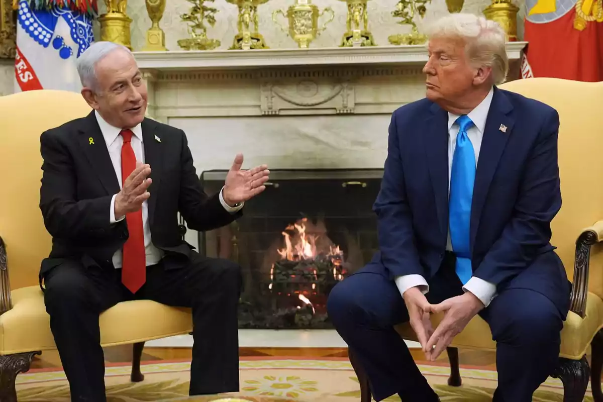 Two men in suits sit facing each other, conversing in front of a lit fireplace in an elegant room. Two men in suits sit facing each other, conversing in front of a lit fireplace in an elegant room.