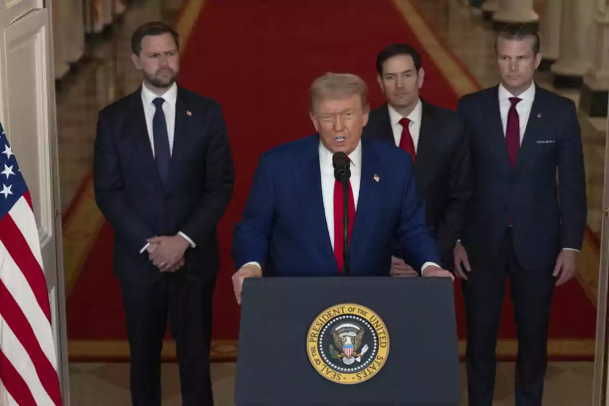 Un hombre de traje azul habla en un podio presidencial mientras tres hombres de traje oscuro lo acompañan al fondo en un salón con alfombra roja y una bandera de Estados Unidos a la izquierda