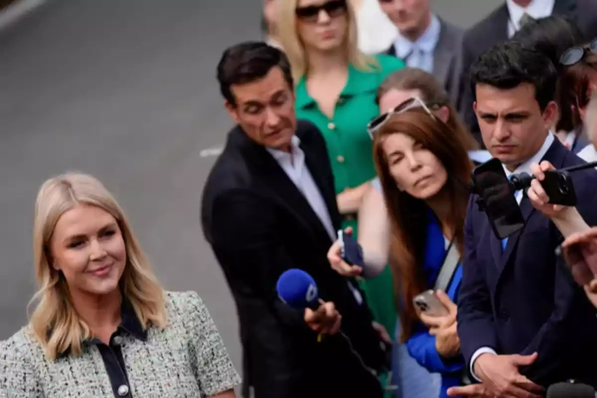 Una mujer rubia sonríe mientras un grupo de periodistas y reporteros la rodea con micrófonos y cámaras. Una mujer rubia sonríe mientras un grupo de periodistas y reporteros la rodea con micrófonos y cámaras.