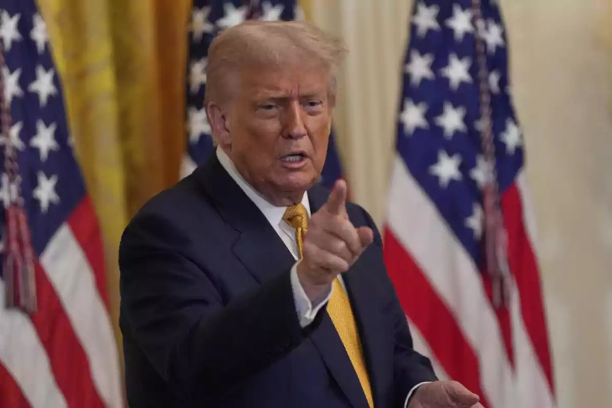 Hombre de traje señalando con el dedo mientras habla, con banderas de Estados Unidos al fondo Hombre de traje señalando con el dedo mientras habla, con banderas de Estados Unidos al fondo
