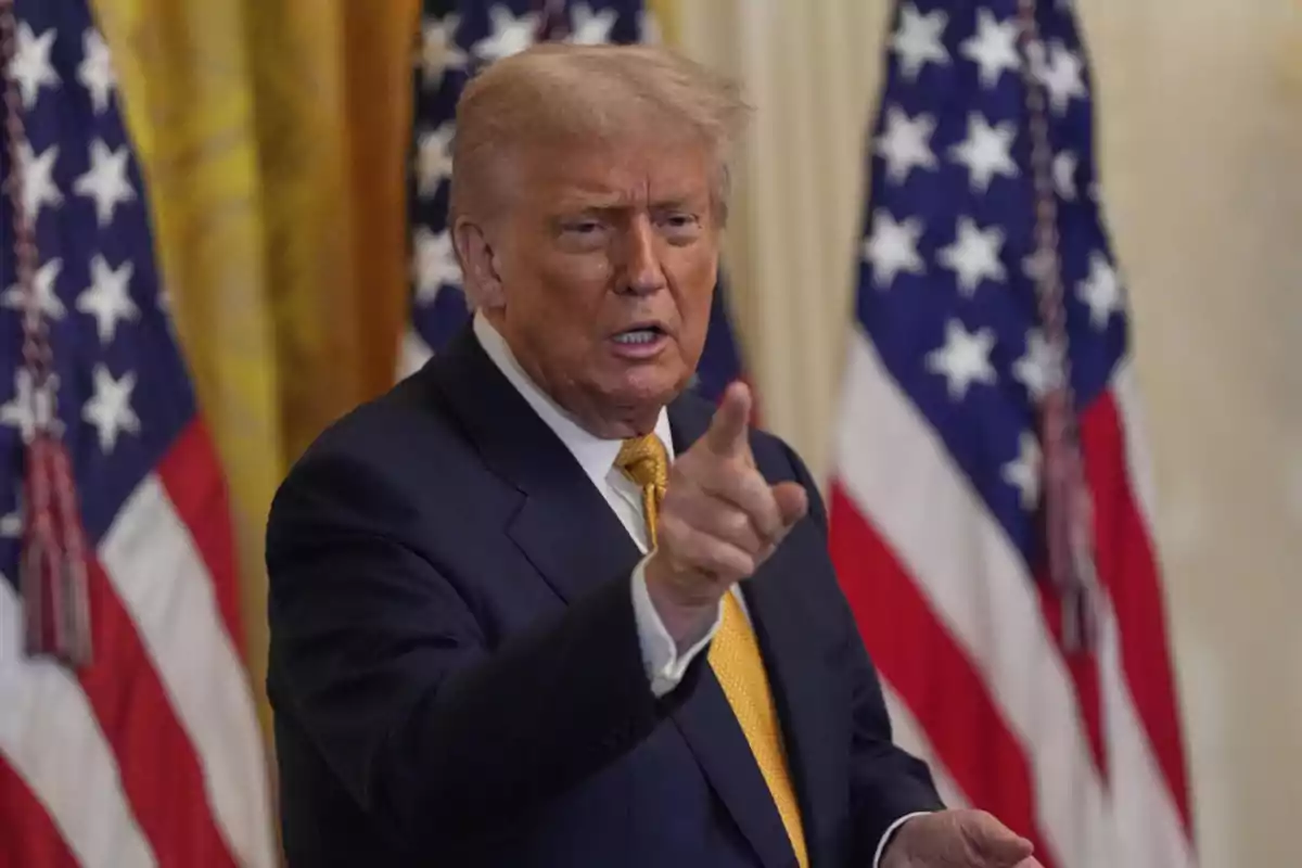 Man in a suit pointing with his finger while speaking, with United States flags in the background Man in a suit pointing with his finger while speaking, with United States flags in the background
