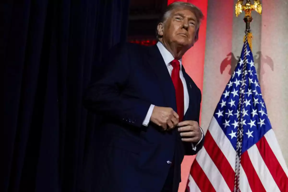 Man in a dark suit and red tie standing next to a United States flag Man in a dark suit and red tie standing next to a United States flag