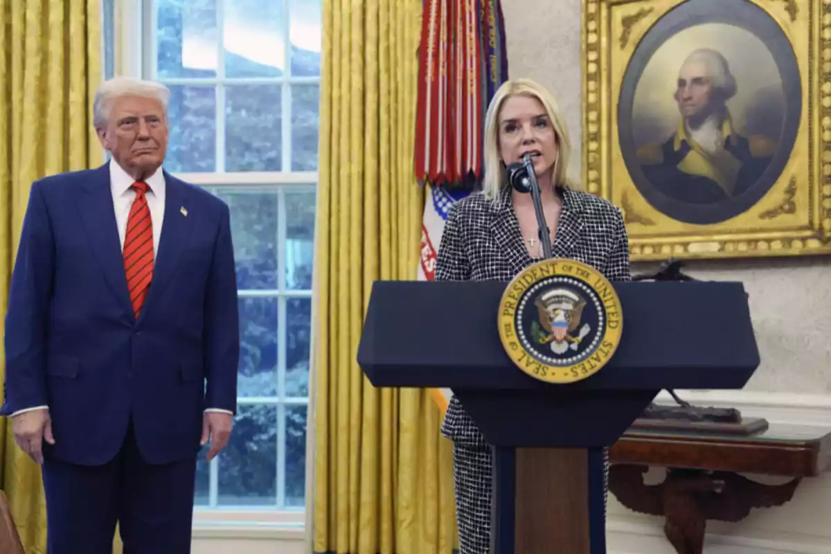 A man in a blue suit and red tie is standing next to a woman who is speaking at a presidential podium in a room with golden curtains and an old portrait on the wall. A man in a blue suit and red tie is standing next to a woman who is speaking at a presidential podium in a room with golden curtains and an old portrait on the wall.